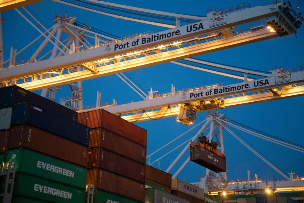 Illuminated cargo cranes at the Port of Baltimore with stacked containers at dusk.