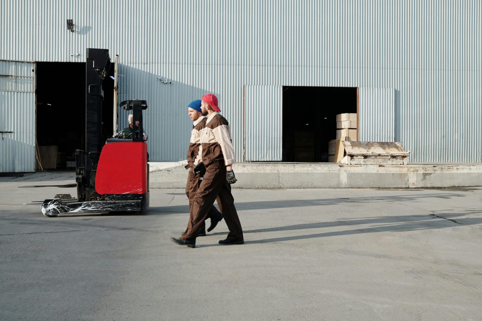 Two workers in uniforms walking outdoors near a forklift at a warehouse.