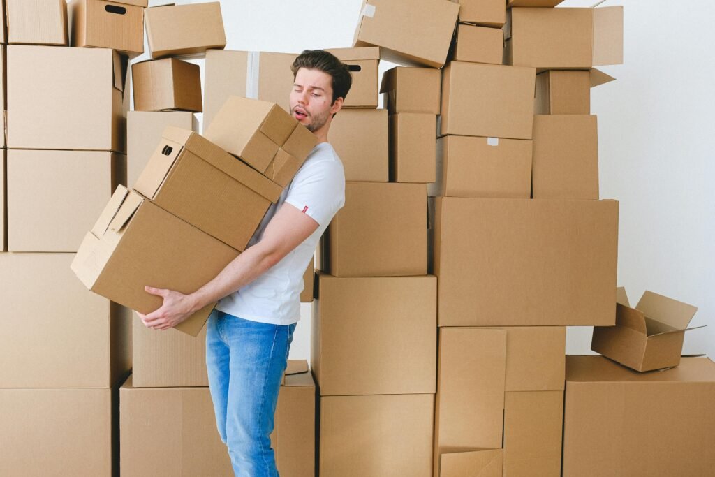 Young man carrying cardboard boxes during a home move, surrounded by a pile of packed containers indoors.