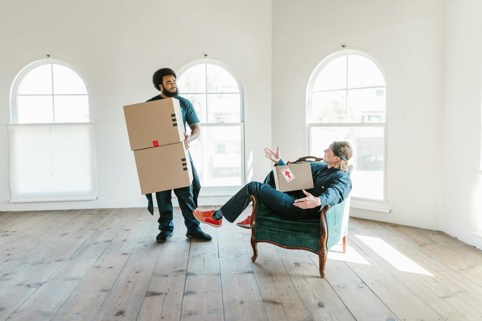 Two men, one sitting and one standing, moving boxes in a bright empty room with arched windows.