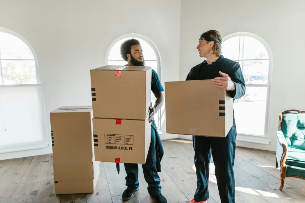 Two professional movers carrying cardboard boxes inside a bright room with large windows.