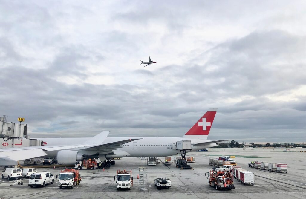 Swiss aircraft parked on Los Angeles airport apron with a plane flying overhead on a cloudy day.